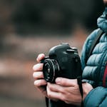 Close-up image of a photographer holding a DSLR camera outdoors in a puffer jacket, showcasing focus and creativity.